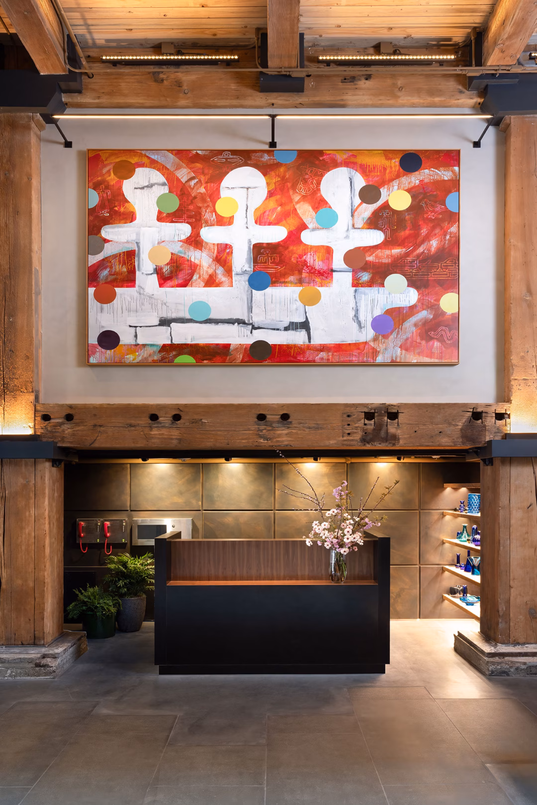 Khách sạn Populus Seattle – Điểm nhấn kiến trúc với cảnh quan sống động ở Pioneer Square A modern reception area with a small dark desk, flower arrangement, wooden beams, wall shelves, and a large abstract painting with red, white, and colorful dot patterns above.