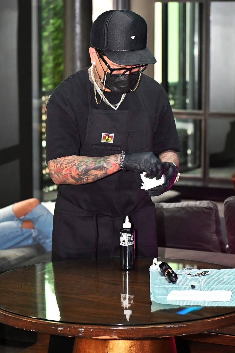A tattoo artist preparing equipment at a studio workspace, wearing gloves and a face mask