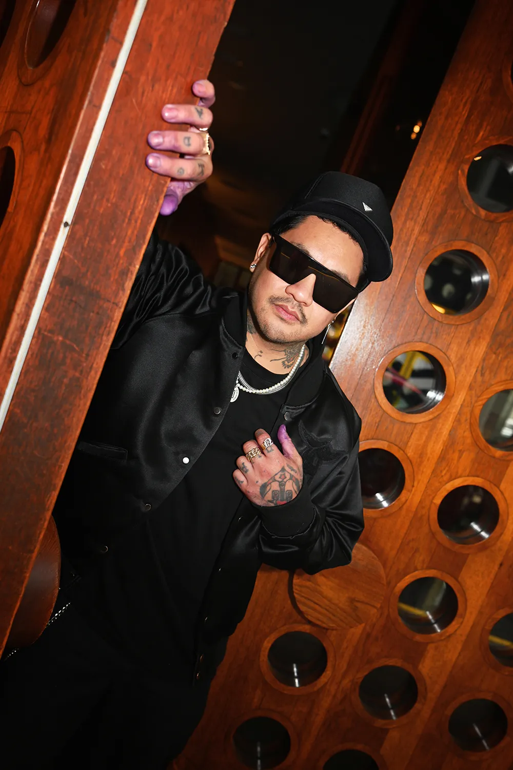 A person in sunglasses and a black jacket standing near a wooden door with circular designs