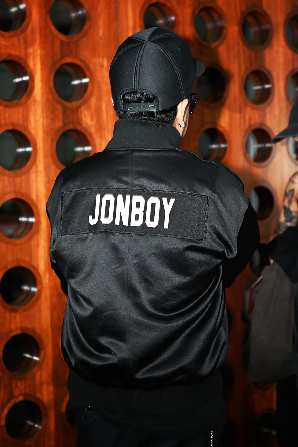 Person with "Jonboy" written on their jacket, standing against a wall with circular cutouts