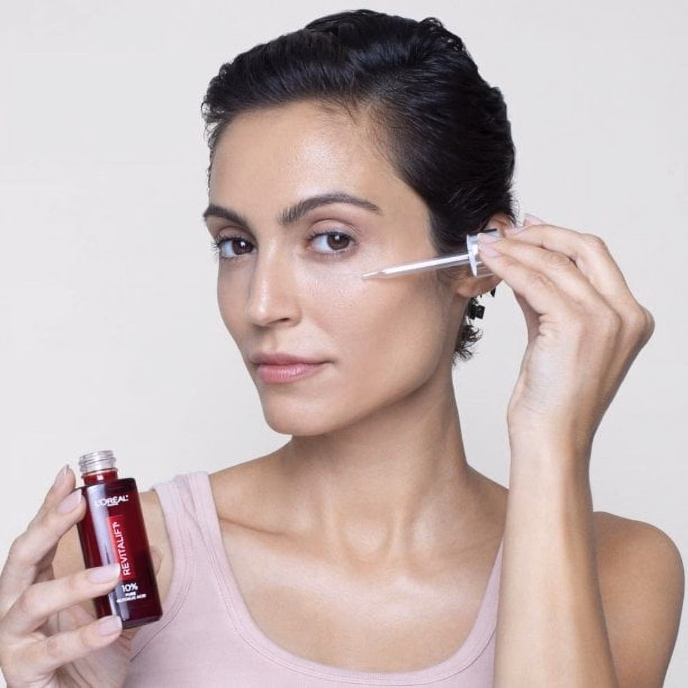 Person applying skincare serum with a dropper, holding a bottle labeled &quot;L'Oreal&quot; in the other hand