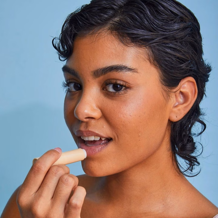 Person applying lip balm to their lips, holding the product in a light-colored tube against a plain background
