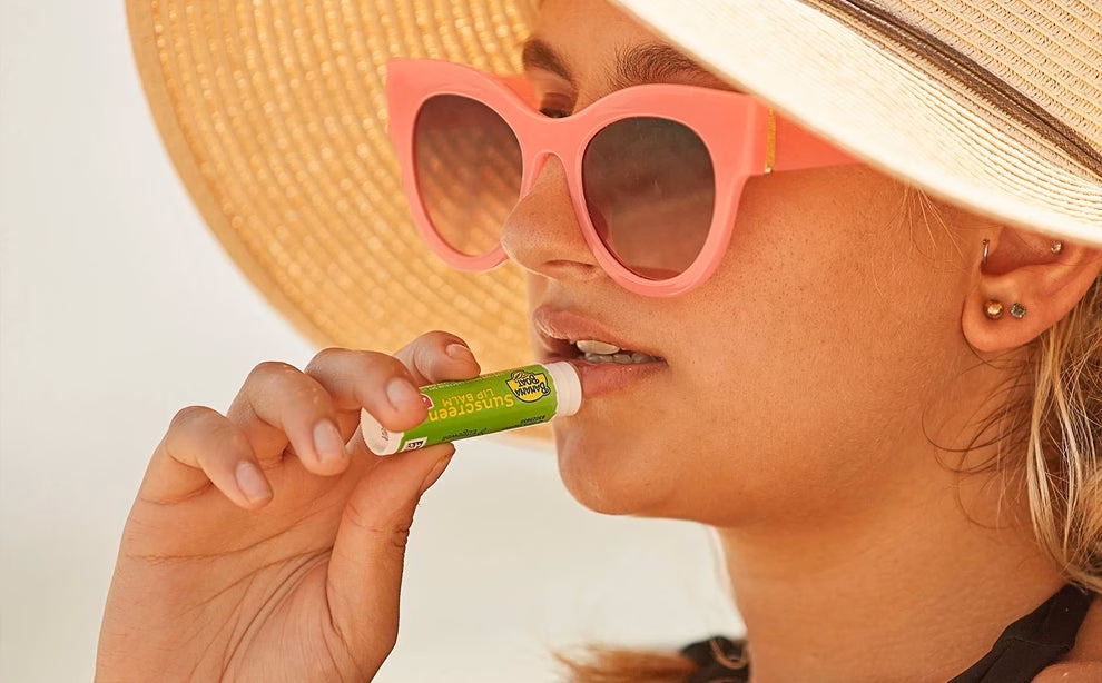 Person wearing a hat and sunglasses applies lip balm. Text: &quot;Soothes dry lips all day. Made with aloe vera &amp;amp; vitamin E.&quot;
