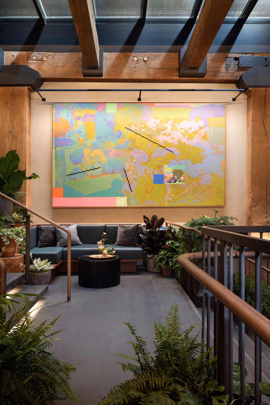 Modern indoor lounge area with a large abstract painting on the wall, surrounded by greenery, wooden beams, and contemporary seating.