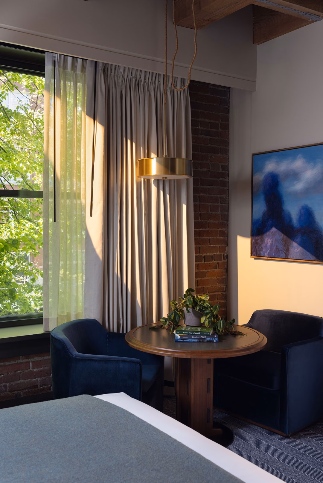 Khách sạn 120 phòng ở Seattle với thiết kế tiền sảnh độc đáo như rừng sống A small round table with two blue chairs sits by a window with sheer curtains. A potted plant and books are on the table, and a painting hangs on an exposed brick wall.