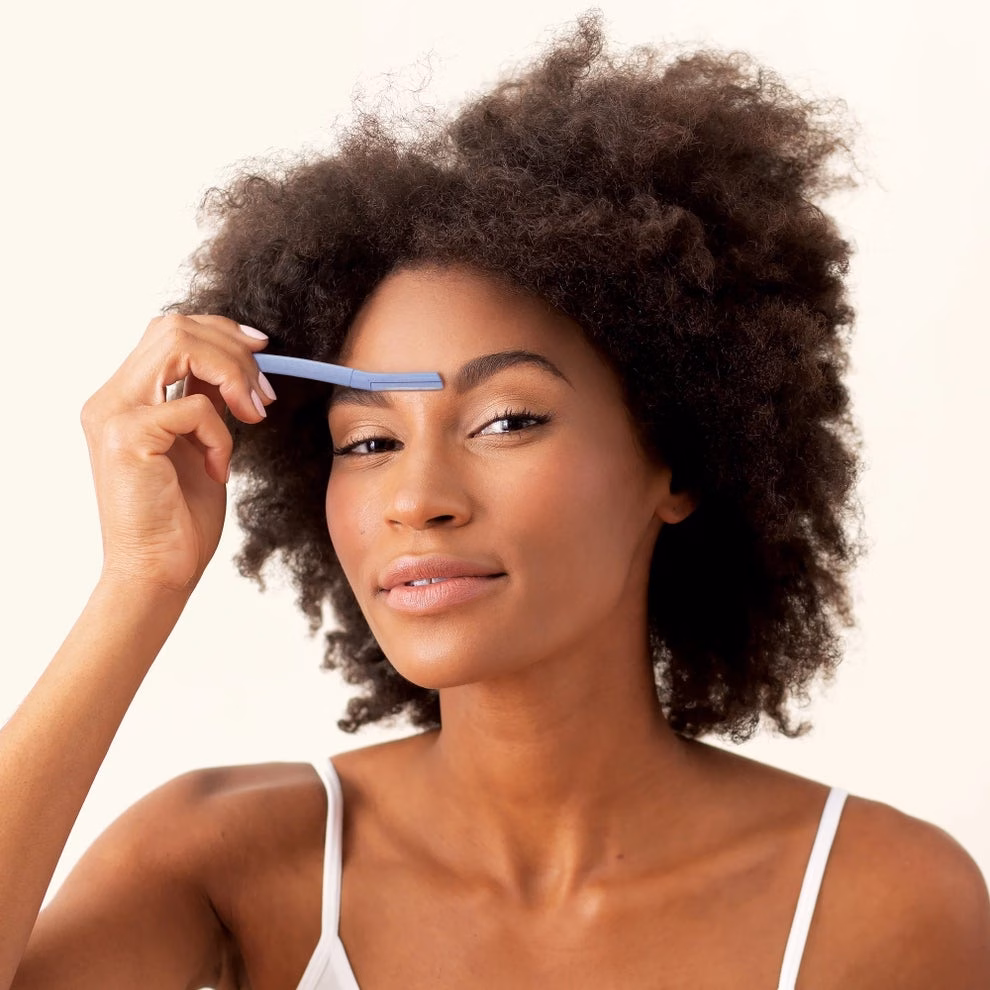 3 sản phẩm chăm sóc da ‘hot’ giúp điều trị mụn, dưỡng ẩm và cải thiện làn da Person using a facial razor on their eyebrow, demonstrating grooming technique. They smile slightly, wearing a thin-strapped top